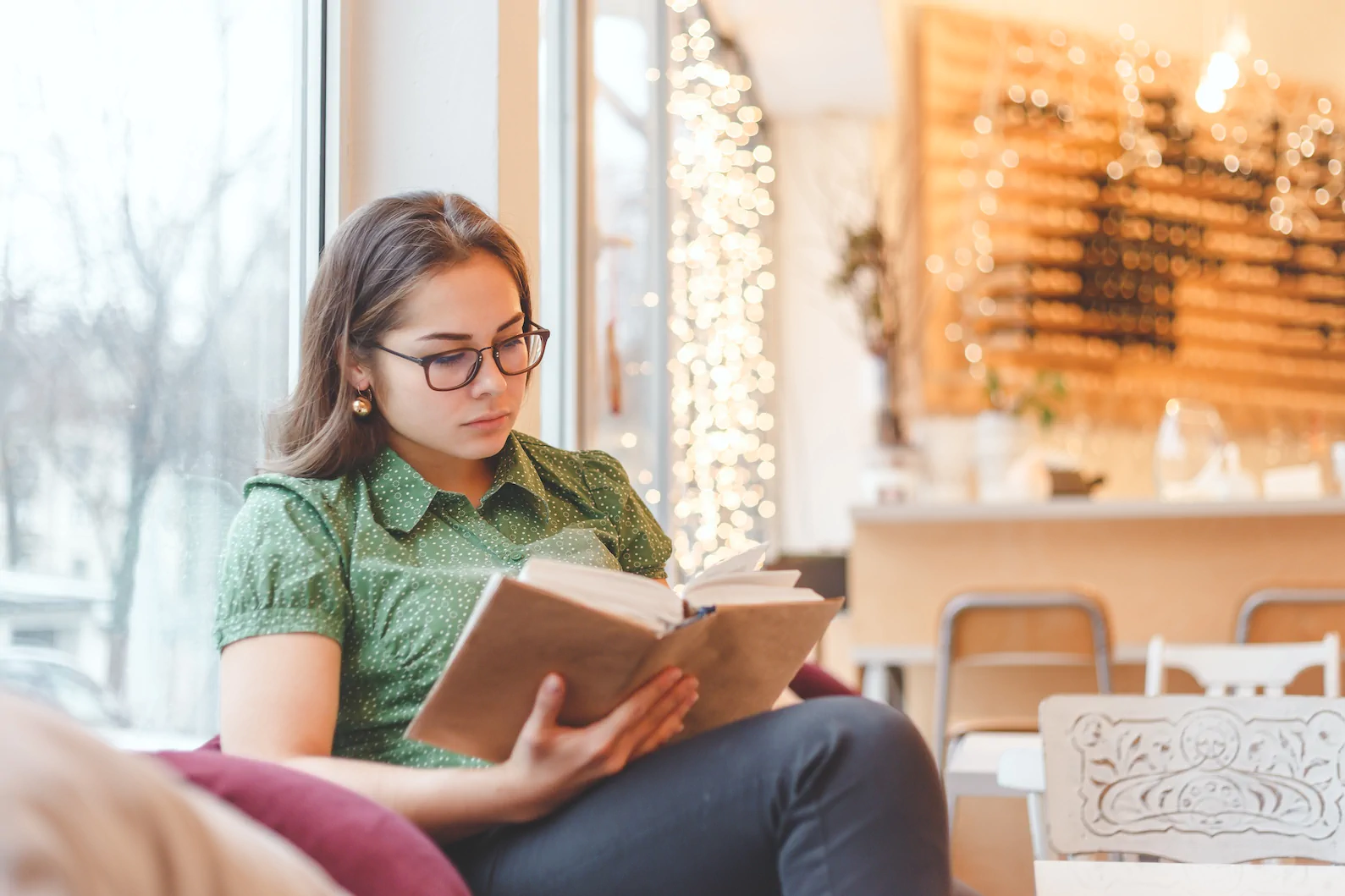 A woman in glasses and a green shirt reads by a cafe window, reminiscent of clients at Always Reddy Nutrition in Pittsburgh, PA, owned by dietitian Allison Reddy specializing in comprehensive nutrition care. in Pennsylvania