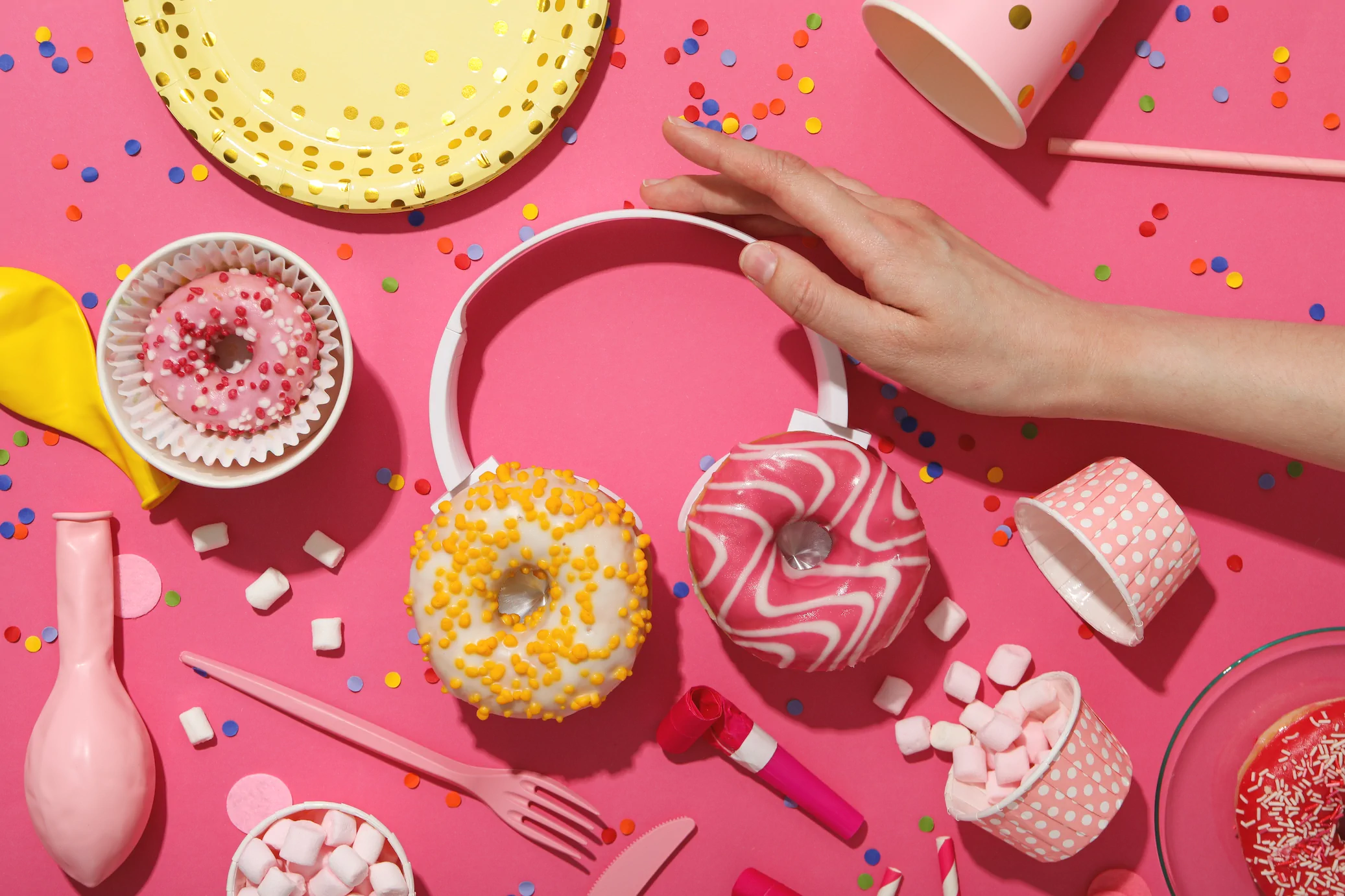 A hand reaches for decorated donuts on headphones amid party supplies, confetti, balloons, cups, and marshmallows on pink—evoking the fun vibe at Always Reddy Nutrition in Pittsburgh, PA. Owned by Allison Reddy—a dietitian specializing in nutrition counseling for neurodivergent people, fertility support, non-diet nutrition and disordered eating. in Pennsylvania