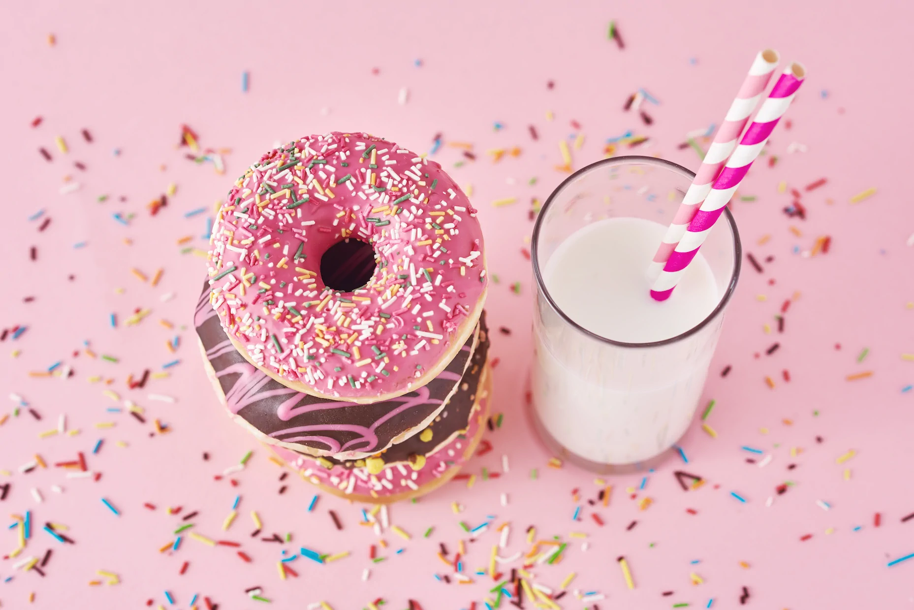 A stack of donuts with pink and chocolate icing beside a glass of milk with pink-striped straws, symbolizing treats enjoyed at Always Reddy Nutrition, Pittsburgh, owned by dietitian Allison Reddy. in Pennsylvania