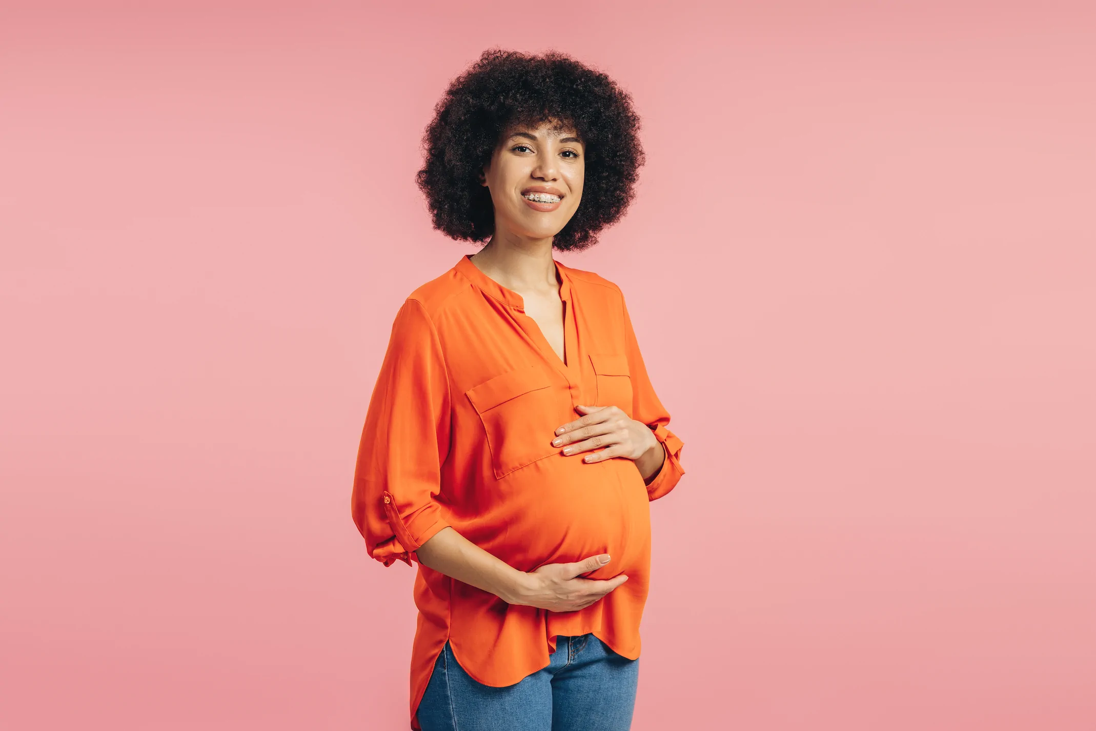 A pregnant woman in an orange blouse and blue jeans stands smiling, holding her belly, against a pink background—reflecting Always Reddy Nutrition in Pittsburgh, PA, owned by Allison Reddy, dietitian for neurodivergent and fertility nutrition. in Pennsylvania