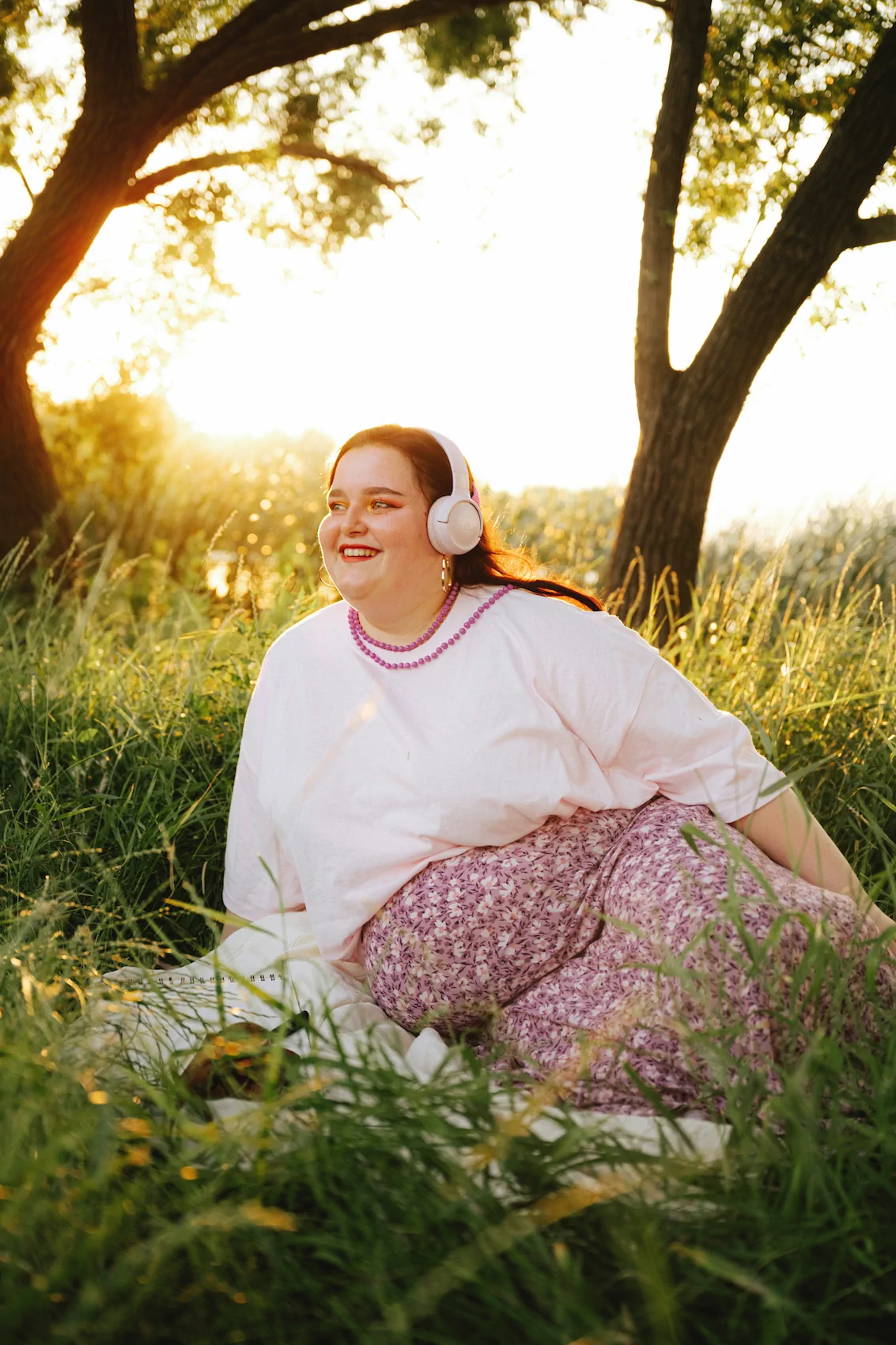 A woman sitting on a blanket in tall grass at sunset, enjoying headphones and smiling, just outside Always Reddy Nutrition in Pittsburgh, PA—owned by dietitian Allison Reddy, specializing in neurodivergent nutrition care and more. in Pennsylvania