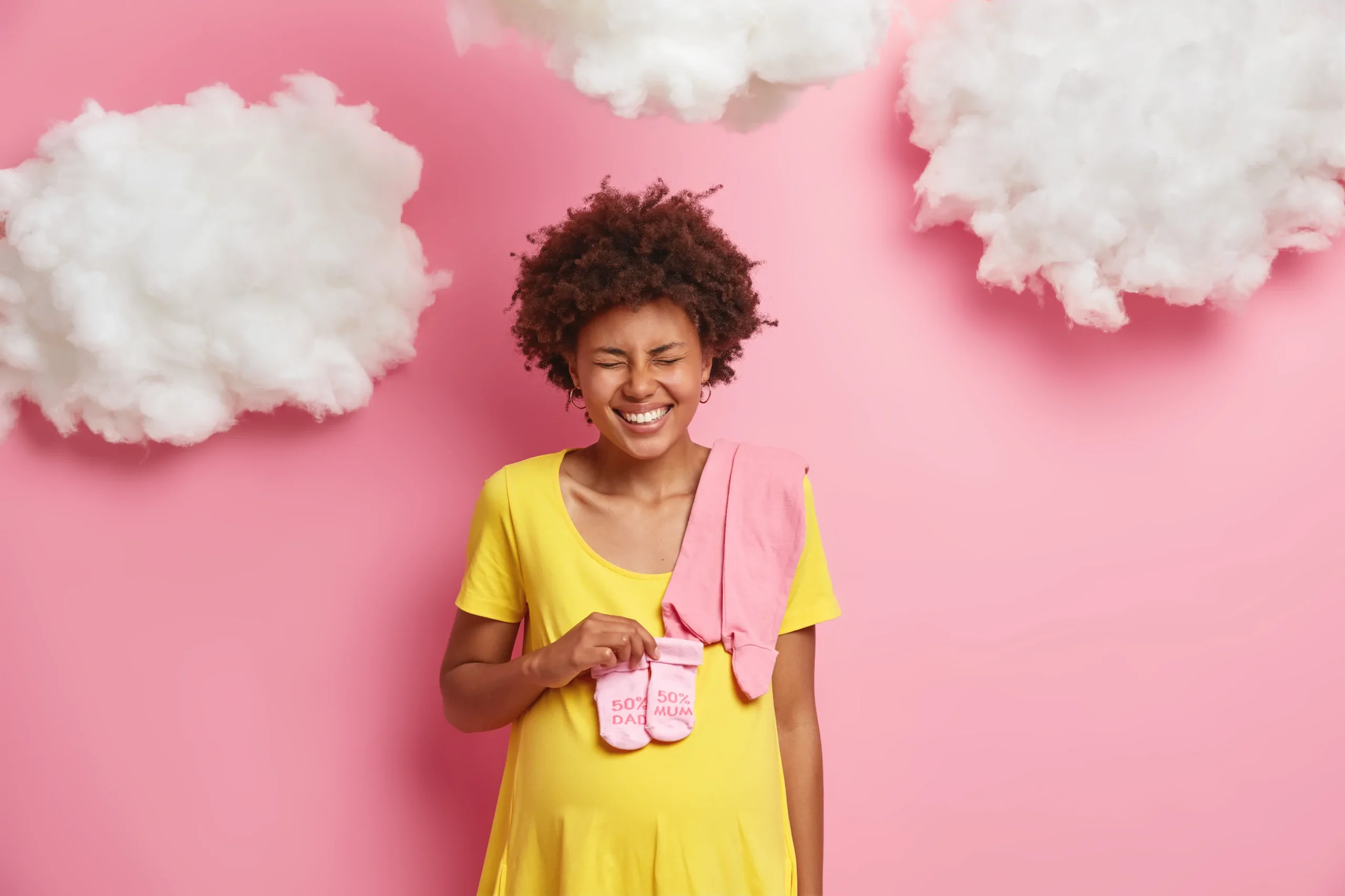 Smiling pregnant woman in yellow dress holds pink baby socks, with a pink towel, against pink clouds; Always Reddy Nutrition in Pittsburgh, PA—owned by dietitian Allison Reddy—offers nutrition for neurodivergent and fertility care. in Pennsylvania